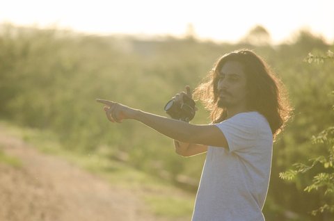 Alex Macedo, fotógrafo de cabelos longos, aponta para o horizonte com uma câmera na mão em contraluz.
